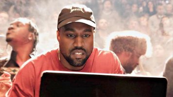 Kanye west with beard, wearing a red t shirt and brown hat in front of computer, book