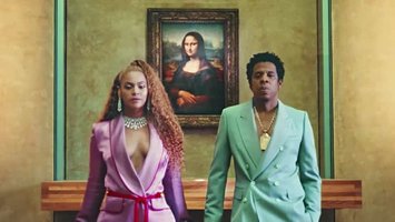 the carters standing in front of Monalisa paint, new album. beyonce with a curly ponytail, is wearing a pink suit, with long earrings and big pearl necklace. Jay z is wearing a grey jacket with a big gold necklace